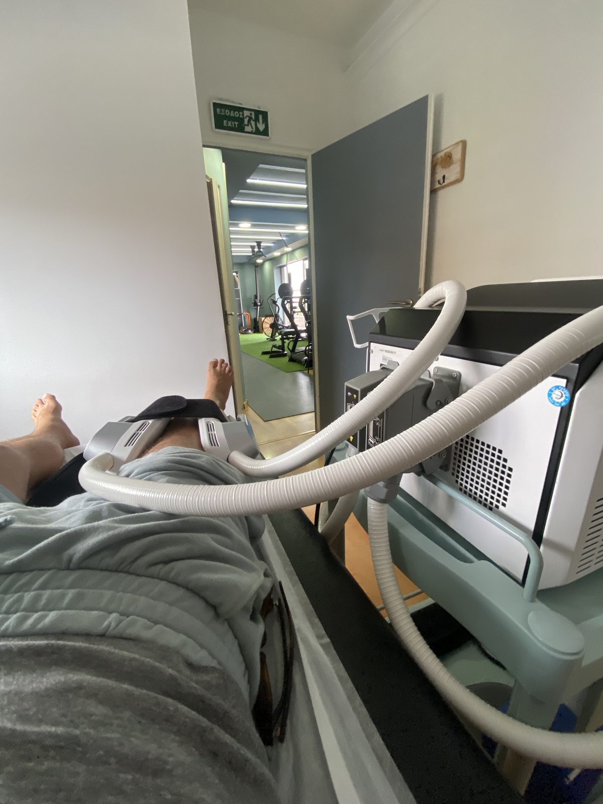 Lying on a physiotherapy machine in a clinic, a Greek exit sign on the wall